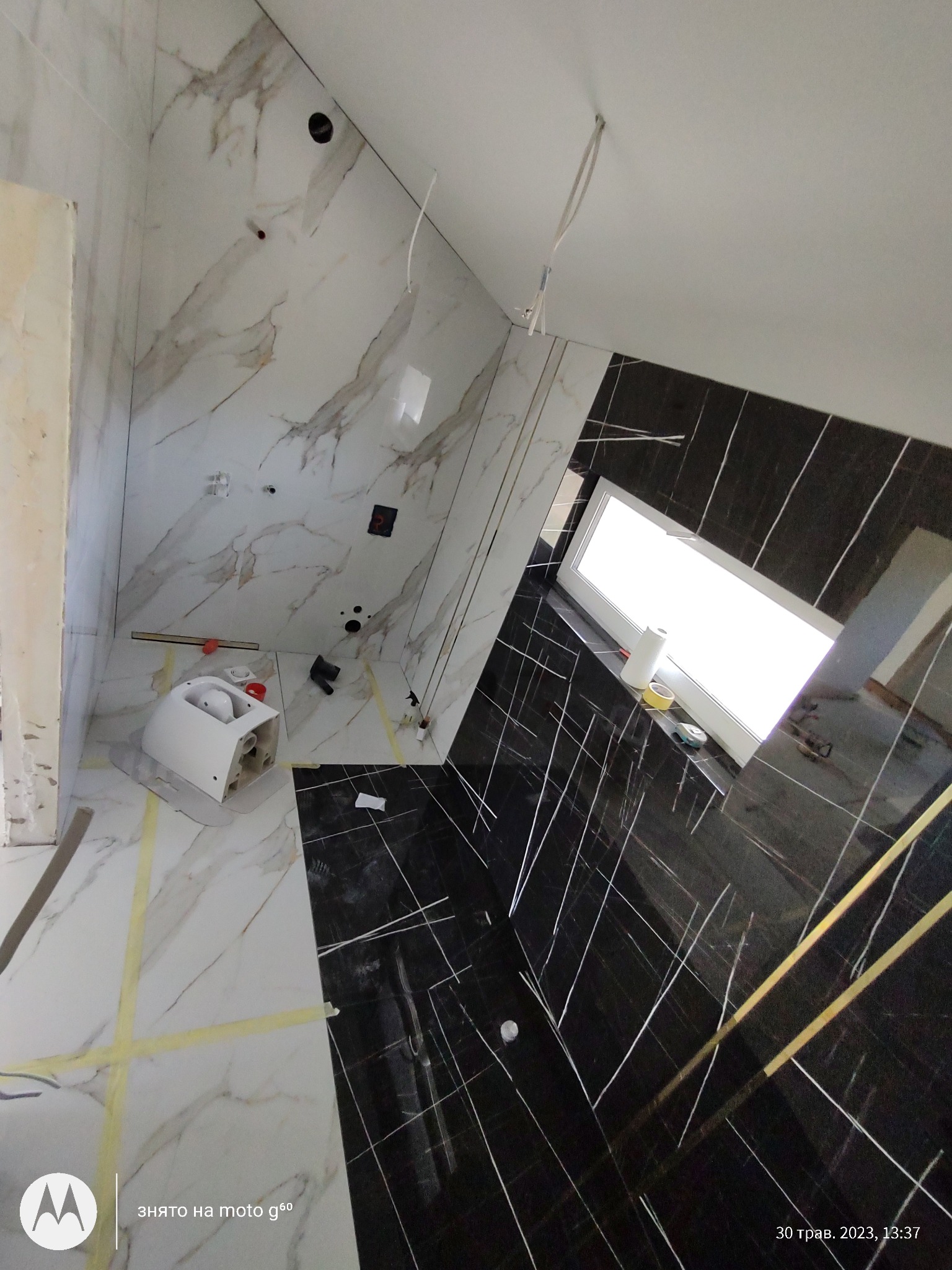 Wnętrze łazienki w trakcie prac wykończeniowych: białe i czarne płytki imitujące marmur, częściowo zamontowana toaleta, widoczne przewody i narzędzia.