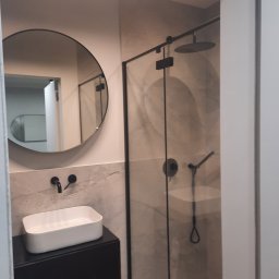 DOMINIK KAHL CRACOW LOCAL TOURS No.1 - Nowoczesna łazienka z okrągłym lustrem, białą umywalką na czarnej szafce i kabiną prysznicową typu walk-in z czarnymi elementami. Minimalistyczny design.