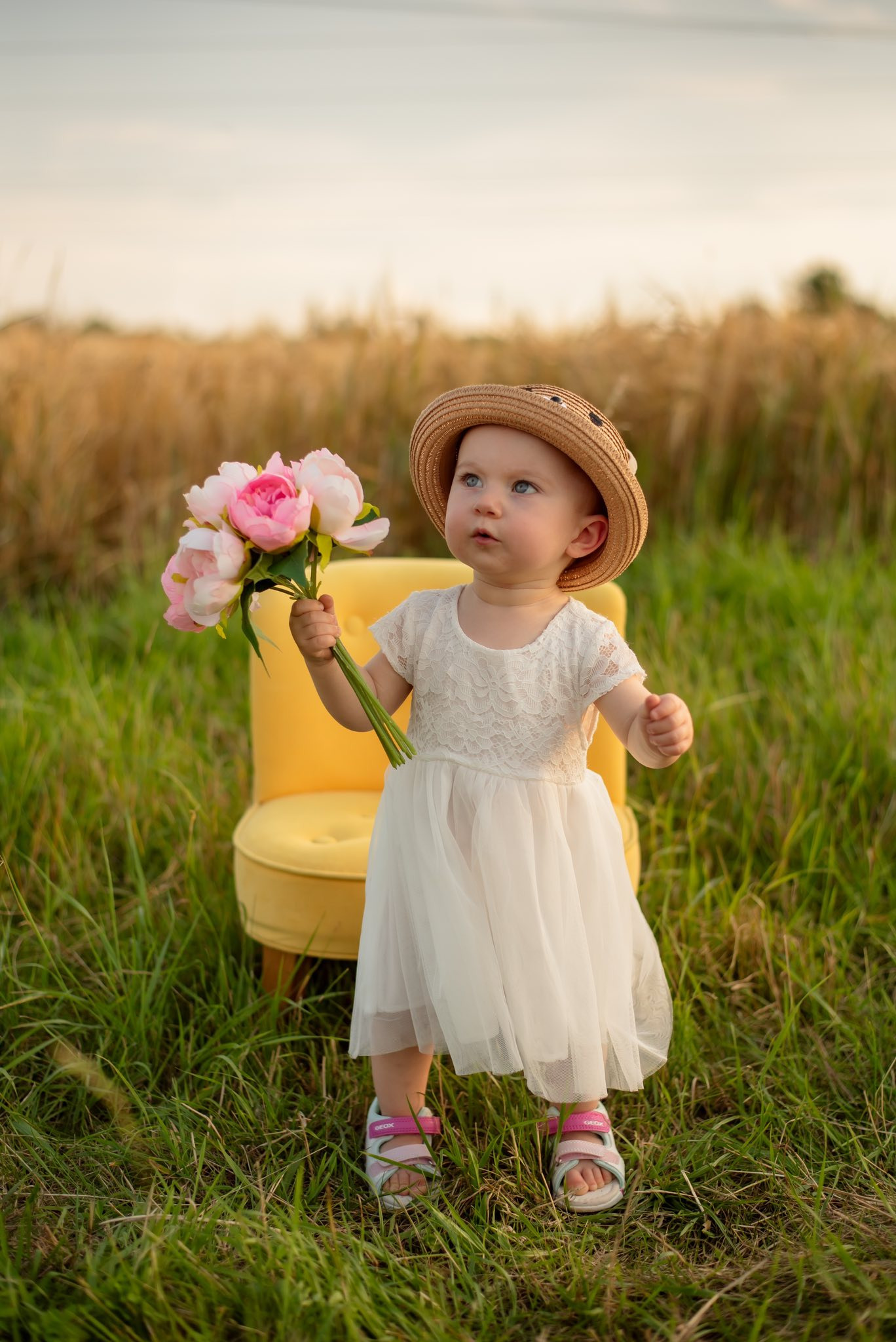 Urocza dziewczynka w białej sukience i kapeluszu trzyma bukiet różowych piwonii, siedząc na żółtym krześle na tle pola pszenicy w ciepłym, naturalnym świetle.