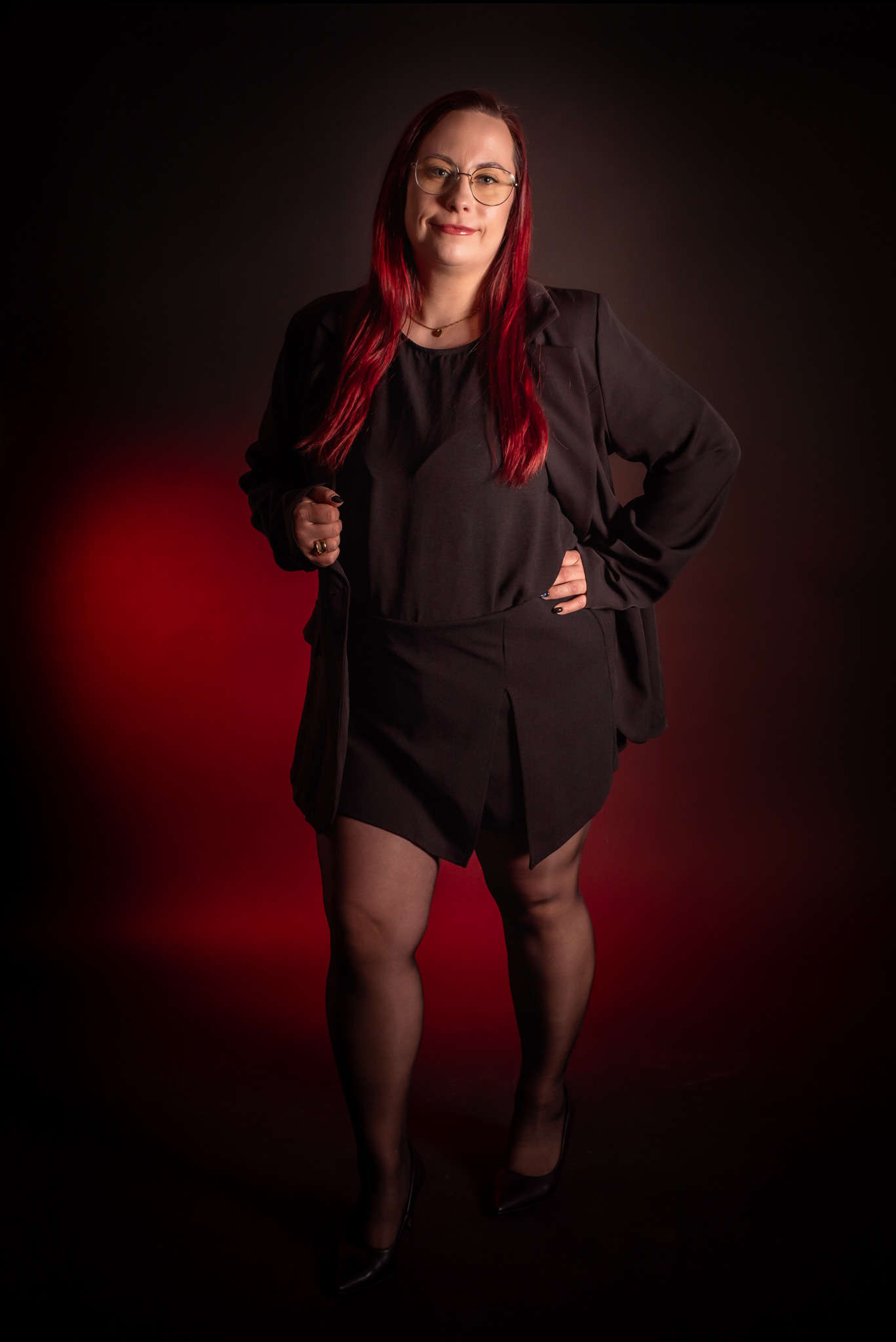 Portret kobiety w okularach, czerwone włosy, czarny strój, pozuje na ciemnym tle z czerwonym akcentem świetlnym. Profesjonalna sesja wizerunkowa.