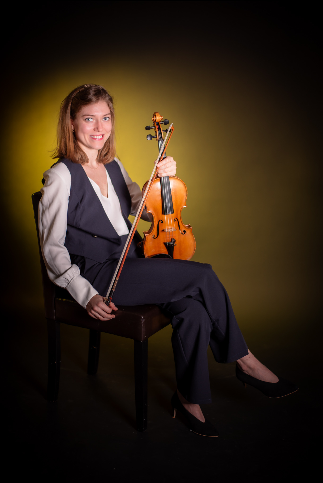 Portret kobiety w garniturze, siedzącej na krześle i trzymającej skrzypce na tle żółto-czarnym. Zdjęcie wykonane w studio w Jaworznie.