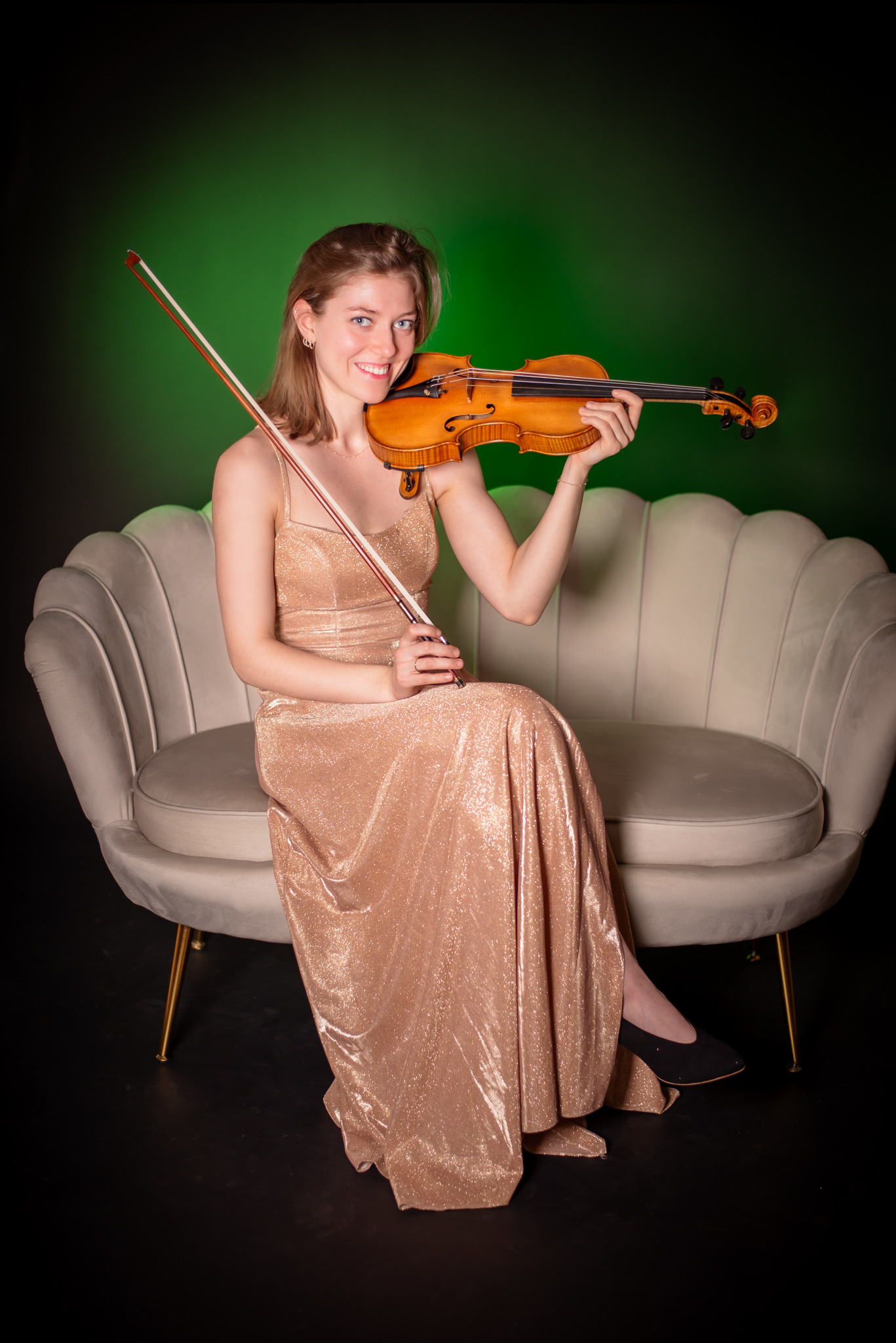 Uśmiechnięta kobieta w złotej sukni siedzi na kanapie z skrzypcami na tle zieleni. Portret w studiu, elegancka sesja z instrumentem.