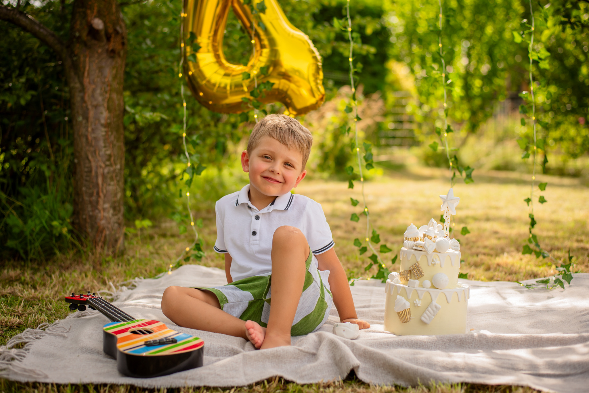 Uśmiechnięty chłopiec siedzi na kocu obok tortu urodzinowego i kolorowej gitary, w tle złoty balon w kształcie cyfry 4 i dekoracje z bluszczu.