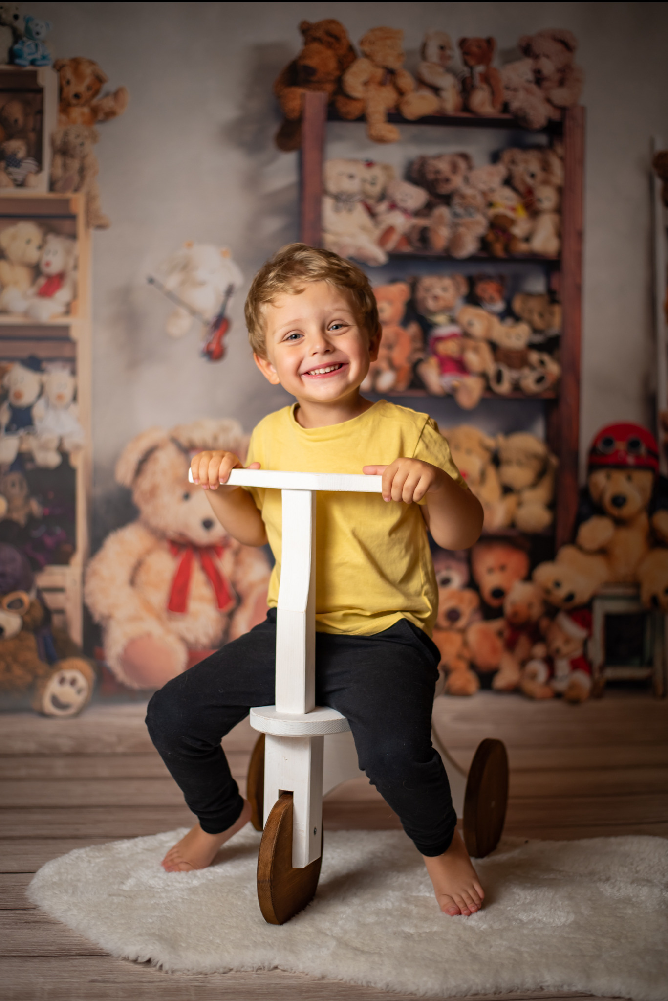 Uśmiechnięty chłopiec w żółtej koszulce siedzi na białym, drewnianym rowerku biegowym na tle ściany z pluszowymi misiami. Radosna fotografia dziecięca.