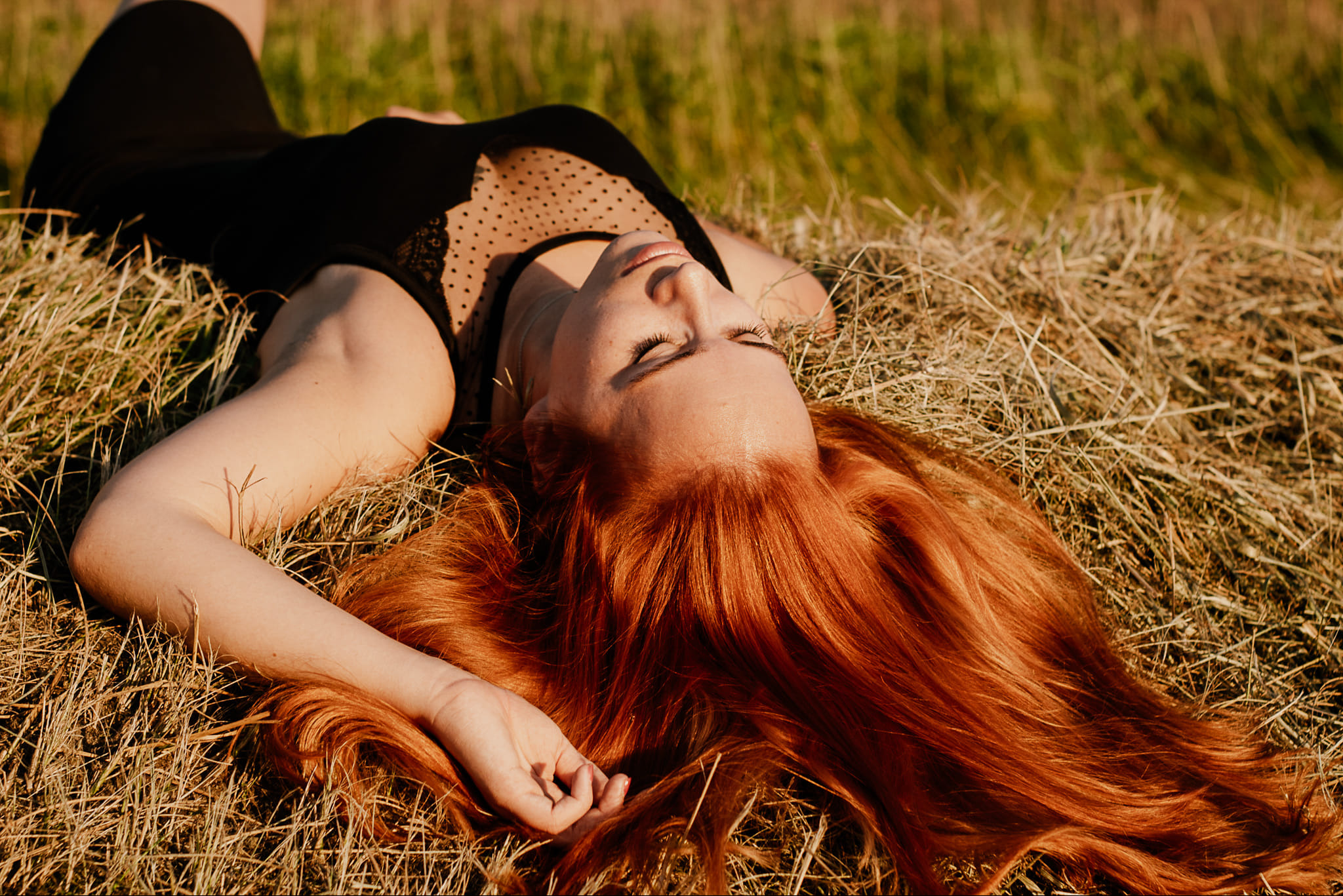 Ruda kobieta w czarnej sukience leży na sianie, z zamkniętymi oczami, w naturalnym świetle. Romantyczna sesja w plenerze.