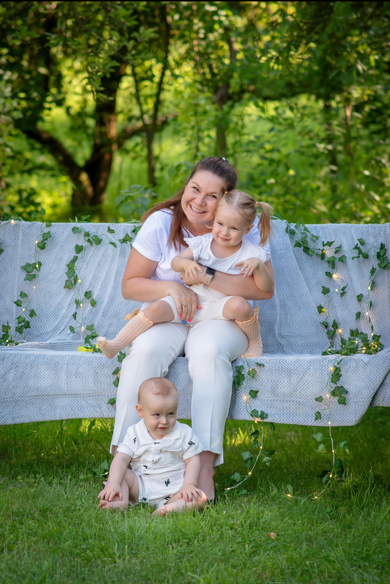 Uśmiechnięta mama z dwiema córkami siedzi na ławce w parku, udekorowanej girlandą z liści i światełek. Na trawie siedzi niemowlę. Sesja rodzinna w plenerze.