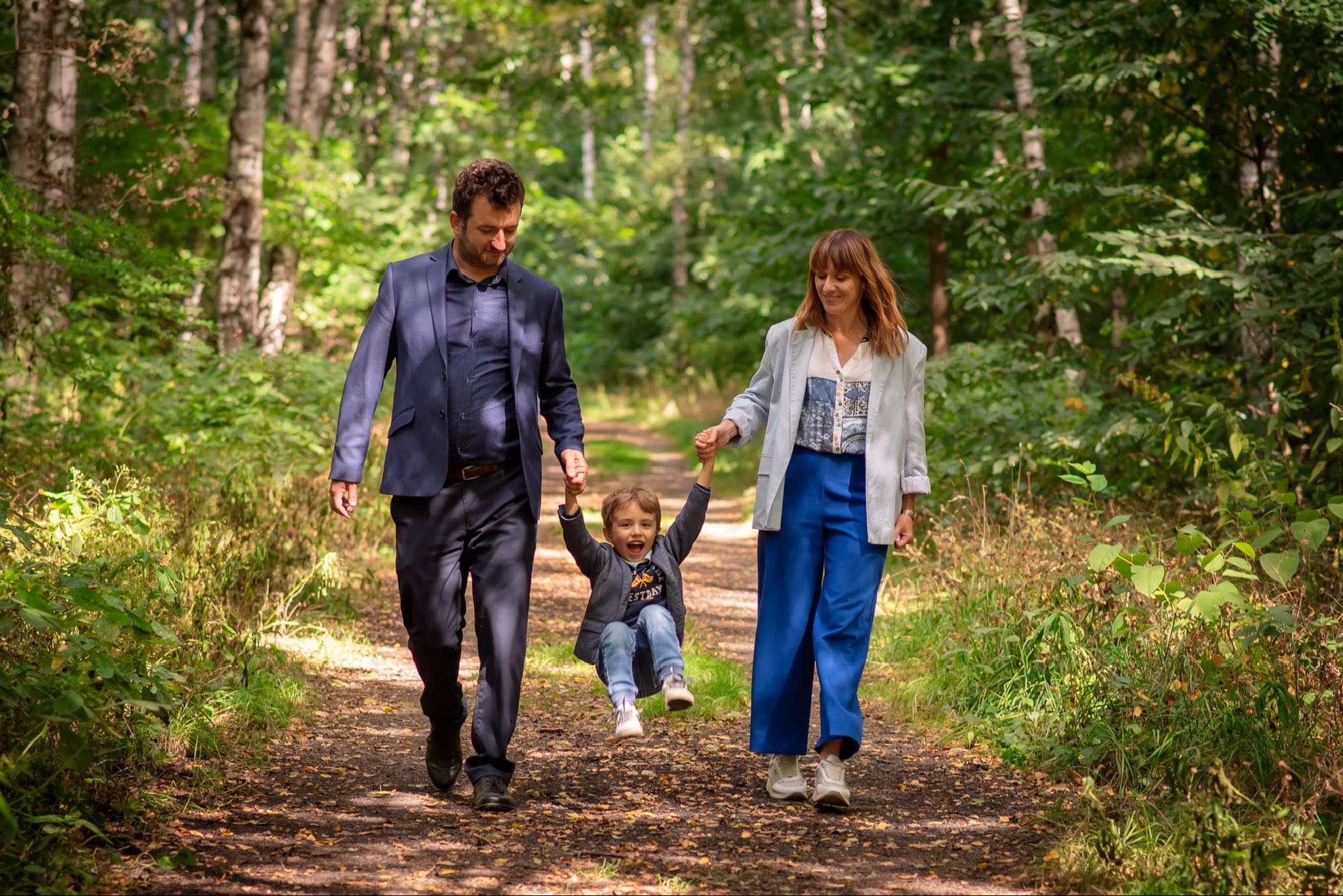 Rodzinne zdjęcie w lesie: rodzice trzymają za ręce uśmiechniętego syna, podnosząc go do góry. Wszyscy ubrani w eleganckie stroje, spacerują leśną ścieżką.