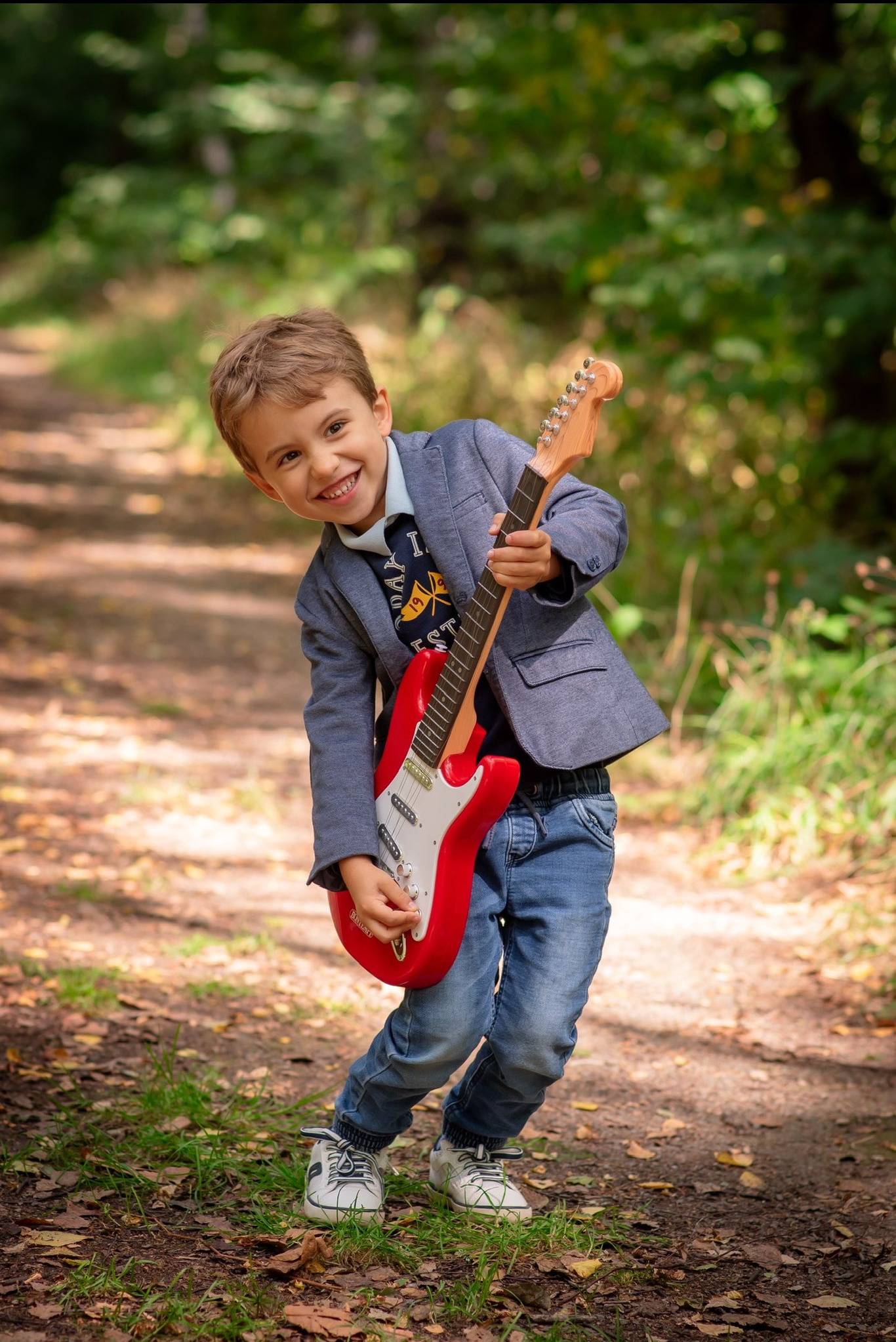 Uśmiechnięty chłopiec w marynarce i jeansach, z czerwoną gitarą elektryczną na leśnej ścieżce, w dynamicznej pozie. Jasne, naturalne światło, zielone tło.