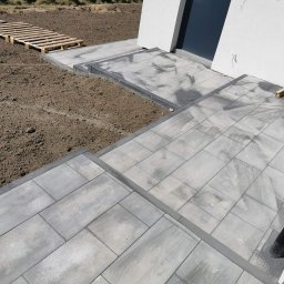 BRUK-BUD JANUSZ KWIDZIŃSKI - Ułożona, szara kostka brukowa przy białym budynku z ciemnym oknem. W tle palety na ziemi i osoba zamiatająca kostkę. Widok z góry.