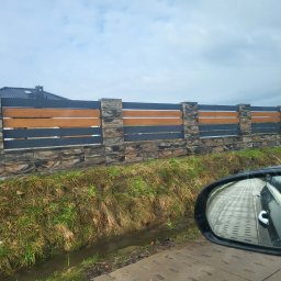 BRUK-BUD JANUSZ KWIDZIŃSKI - Nowoczesne ogrodzenie panelowe z elementami imitującymi kamień i drewno, widoczne z perspektywy samochodu, z odbiciem kostki brukowej w lusterku.