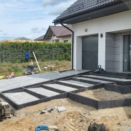 BRUK-BUD JANUSZ KWIDZIŃSKI - Etap prac: układanie kostki brukowej na podjeździe i schodach wejściowych do domu. Widoczne narzędzia i materiały budowlane. Prace brukarskie w toku.