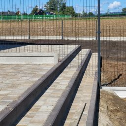 BRUK-BUD JANUSZ KWIDZIŃSKI - Schody z kostki brukowej z obrzeżami, prowadzące do metalowego ogrodzenia panelowego na tle zieleni i błękitnego nieba. Widoczne elementy krajobrazu.