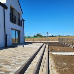 BRUK-BUD JANUSZ KWIDZIŃSKI - Nowoczesny dom z wybrukowanym tarasem i ogrodzeniem panelowym na tle błękitnego nieba. Widoczne schody z kostki brukowej i przygotowany teren pod trawnik.