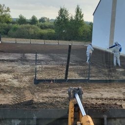 BRUK-BUD JANUSZ KWIDZIŃSKI - Niwelacja terenu pod budowę domu jednorodzinnego. Widoczny operator koparki, pracownicy wyrównujący teren, ogrodzenie panelowe i budynek w tle.