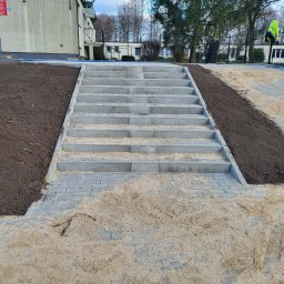 BRUK-BUD JANUSZ KWIDZIŃSKI - Schody z kostki brukowej prowadzące w górę, otoczone ziemią i piaskiem. Widoczne budynki w tle i drzewa. Ujęcie z dołu, podkreślające perspektywę schodów.