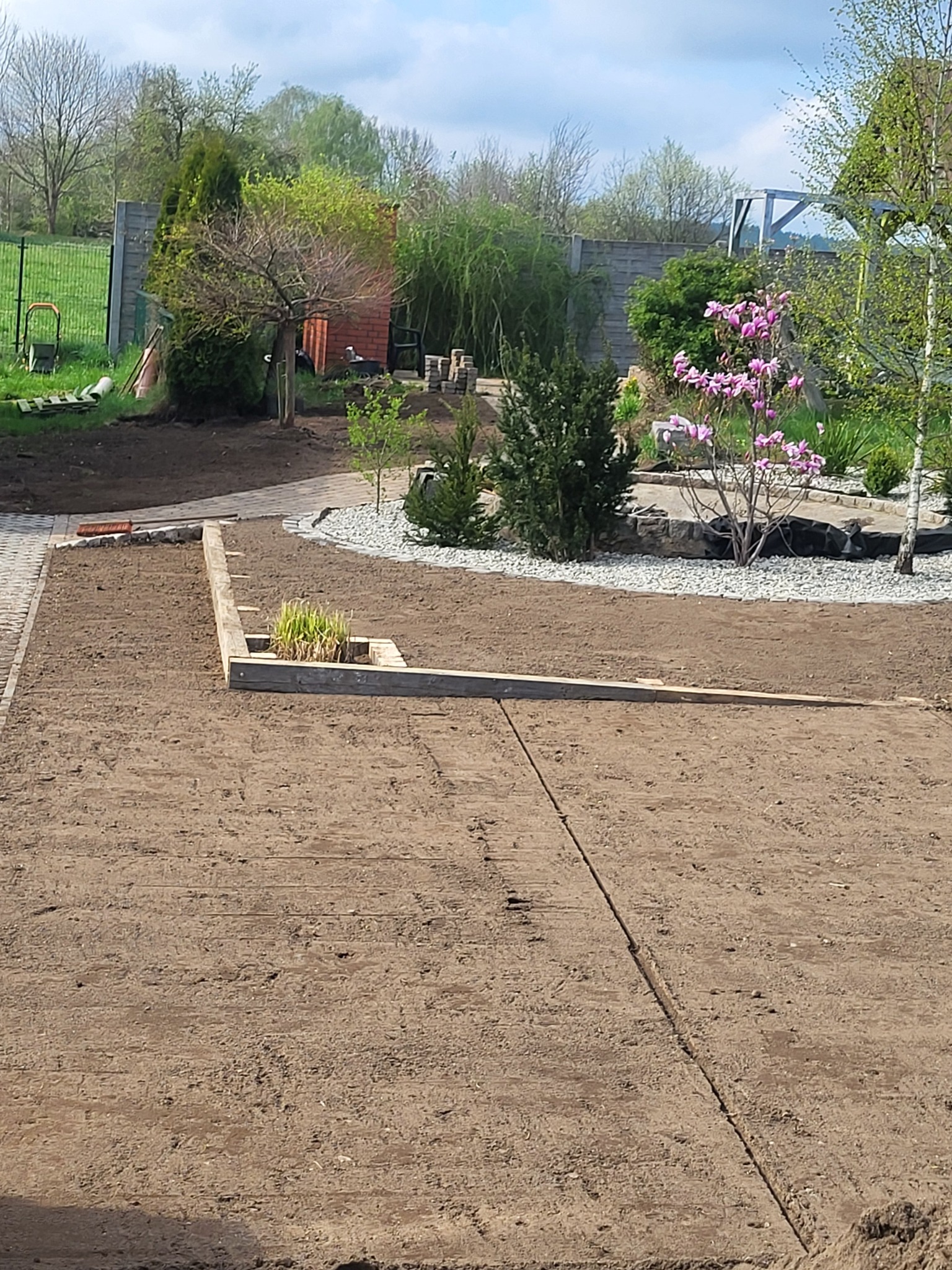 Przygotowany teren pod trawnik z drewnianą obudową rabaty, widoczne ślady po niwelacji, w tle elementy ogrodu: drzewa, krzewy, ogrodzenie panelowe.