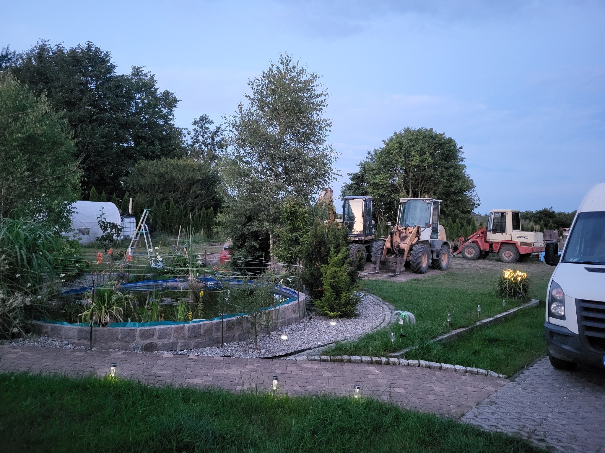 Ogród z oczkiem wodnym obok ścieżki z kostki, w tle maszyny budowlane i biały bus. Wieczorny krajobraz z oświetleniem.