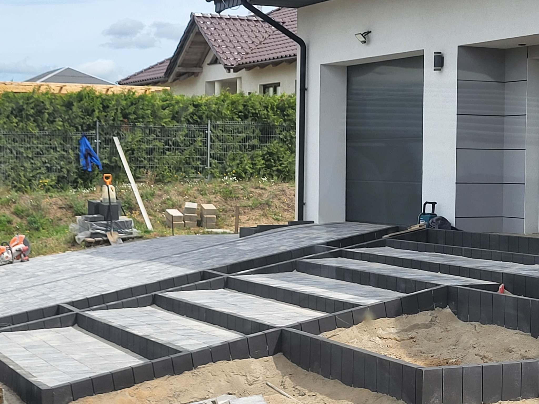 Etap układania kostki brukowej na podjeździe do garażu. Widoczne przygotowane ramy z kostki i piasek. W tle dom z szarą bramą garażową i narzędzia.