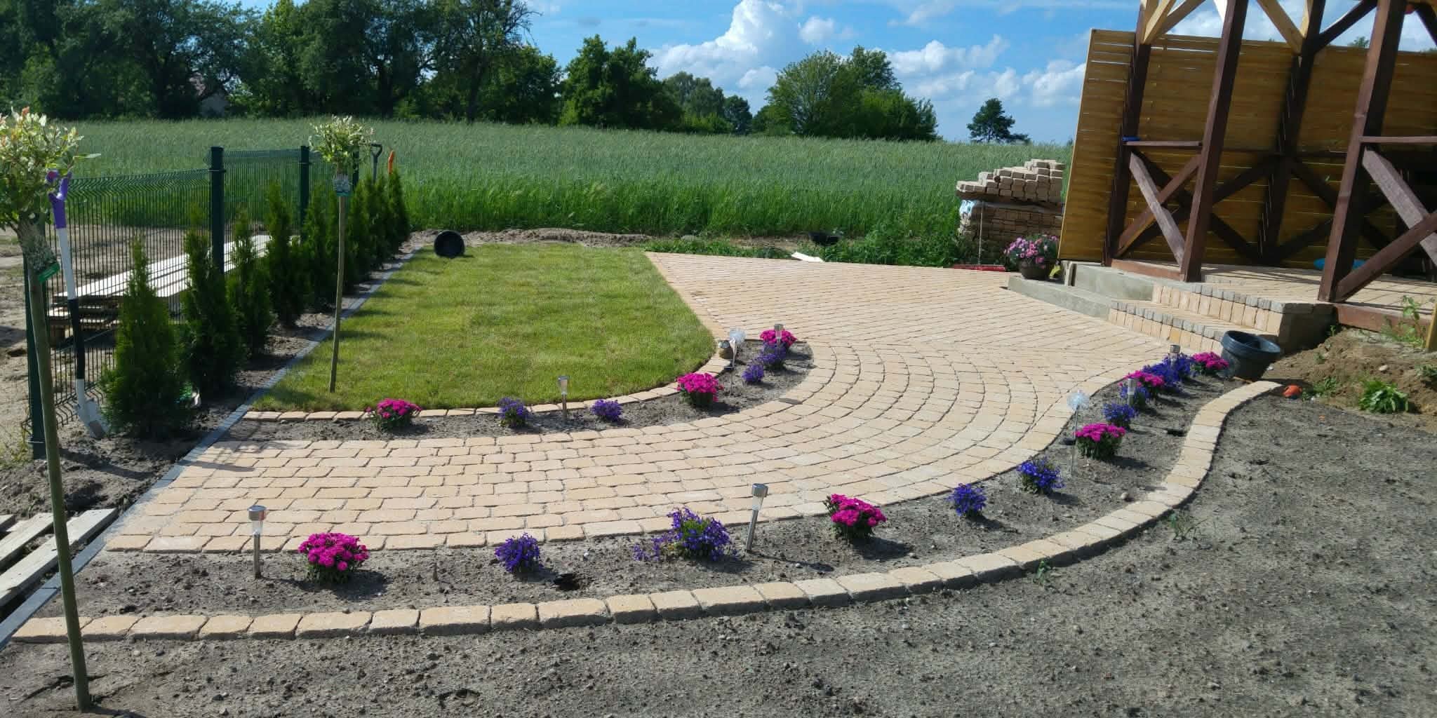 Kremowa kostka brukowa ułożona w łukowaty wzór, obrzeża z kwiatami, trawnik i drewniana altana w tle, oświetlenie solarne wzdłuż ścieżki, słoneczny dzień.