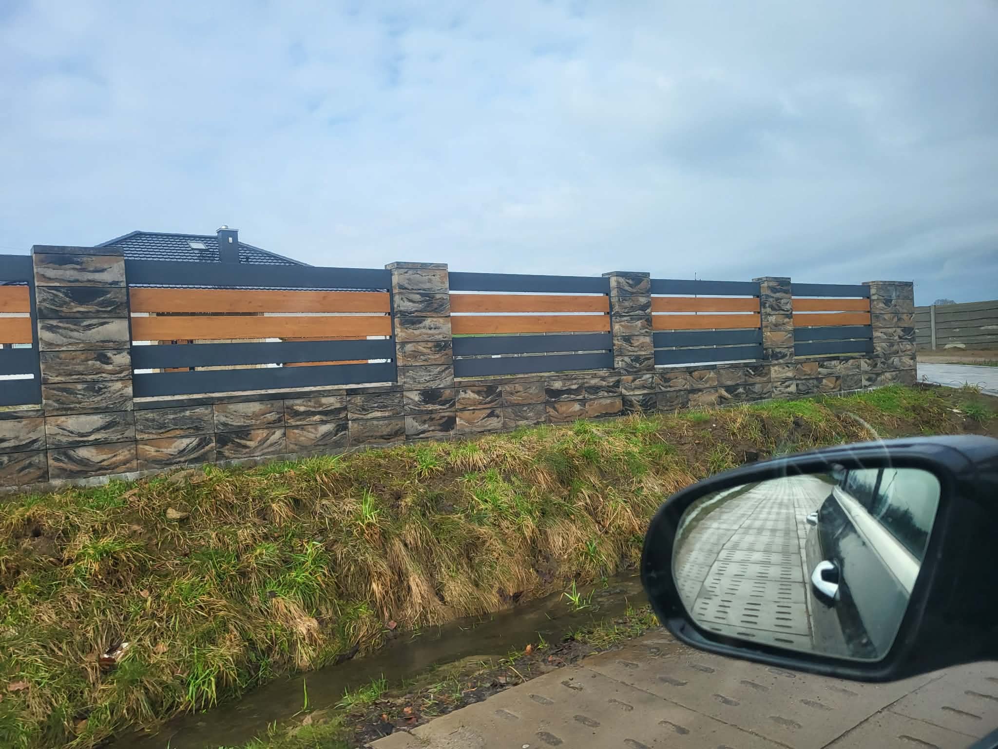 Nowoczesne ogrodzenie panelowe z elementami imitującymi kamień i drewno, widoczne z perspektywy samochodu, z odbiciem kostki brukowej w lusterku.