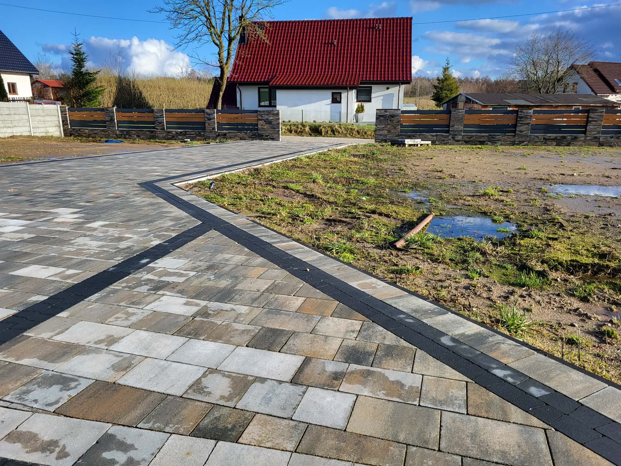 Ułożona kostka brukowa w odcieniach szarości i brązu tworzy ścieżkę w kierunku domu z czerwonym dachem. Widoczny fragment ogrodzenia z elementami drewnianymi.