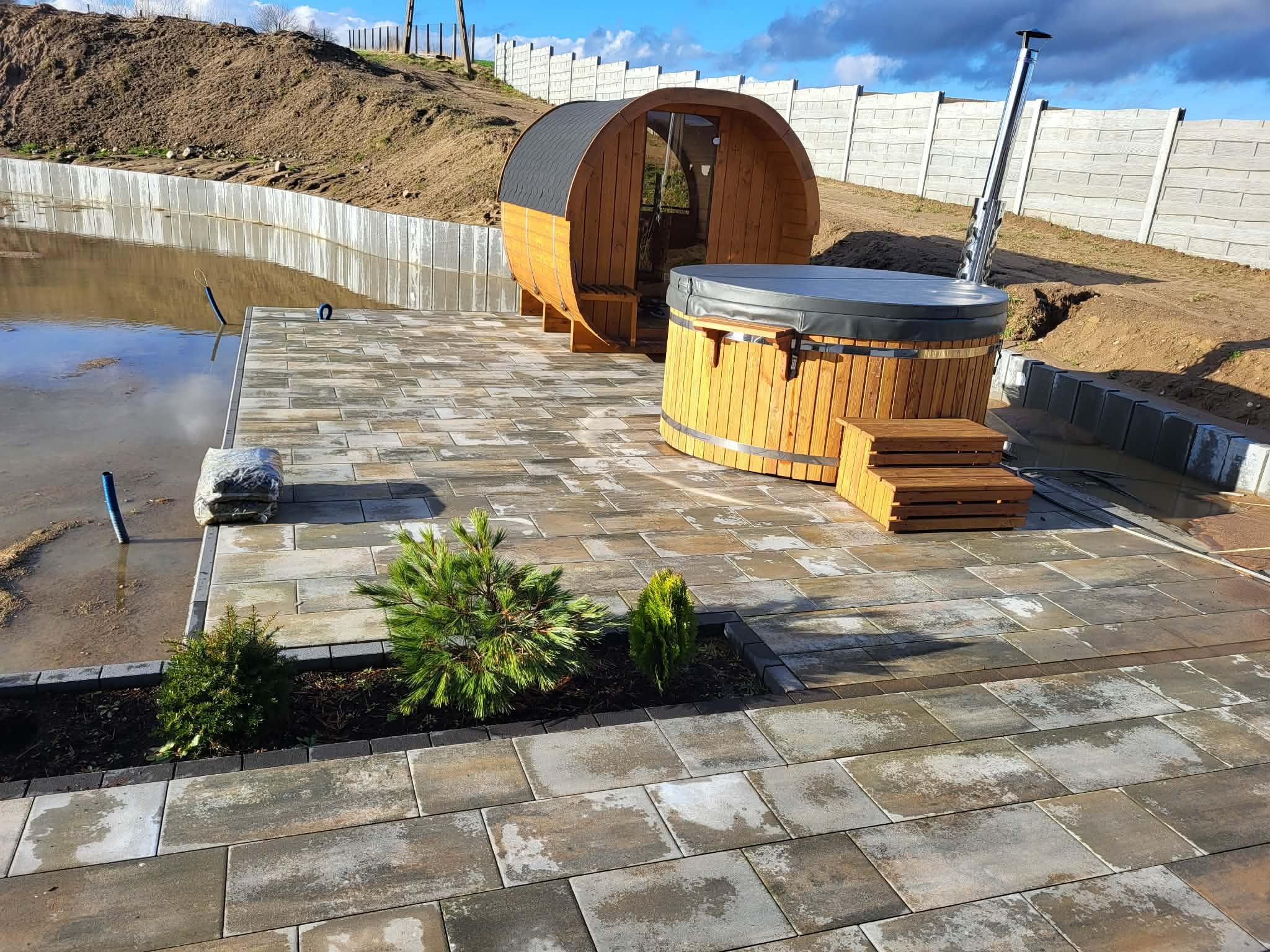 Ułożona kostka brukowa tworząca taras z sauną beczką i jacuzzi. W tle widoczny zbiornik wodny i ogrodzenie panelowe. Całość w naturalnej kolorystyce.