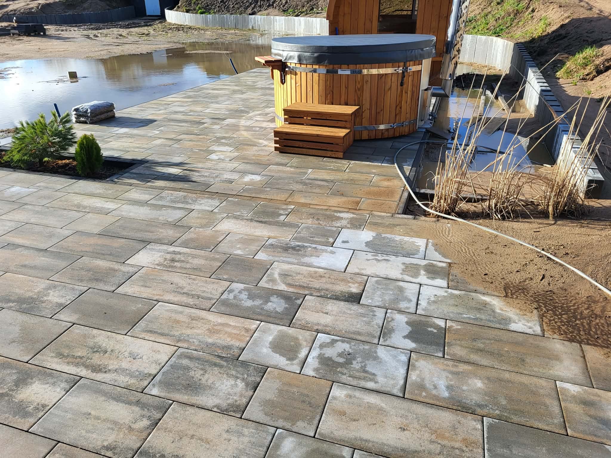 Ułożona kostka brukowa w odcieniach szarości i beżu, prowadząca do drewnianej balii z schodkami. W tle woda i elementy krajobrazu.