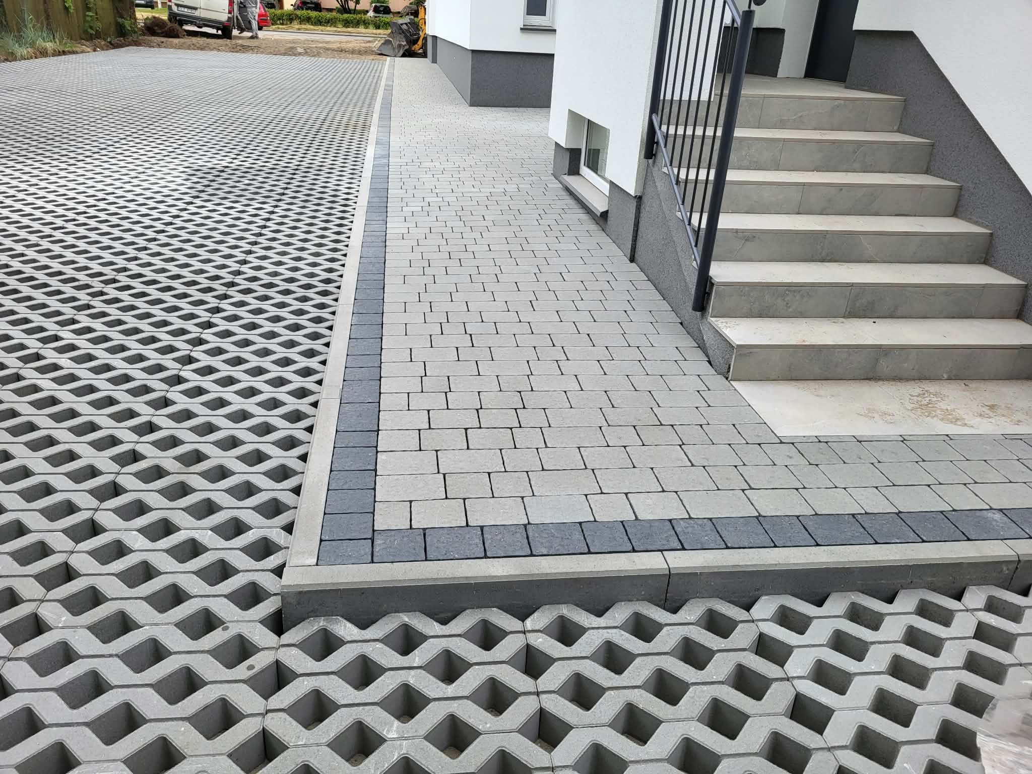 Nowoczesne podejście z kostki brukowej i ażurowych płyt betonowych prowadzące do schodów wejściowych budynku. Widoczny fragment terenu z koparką.