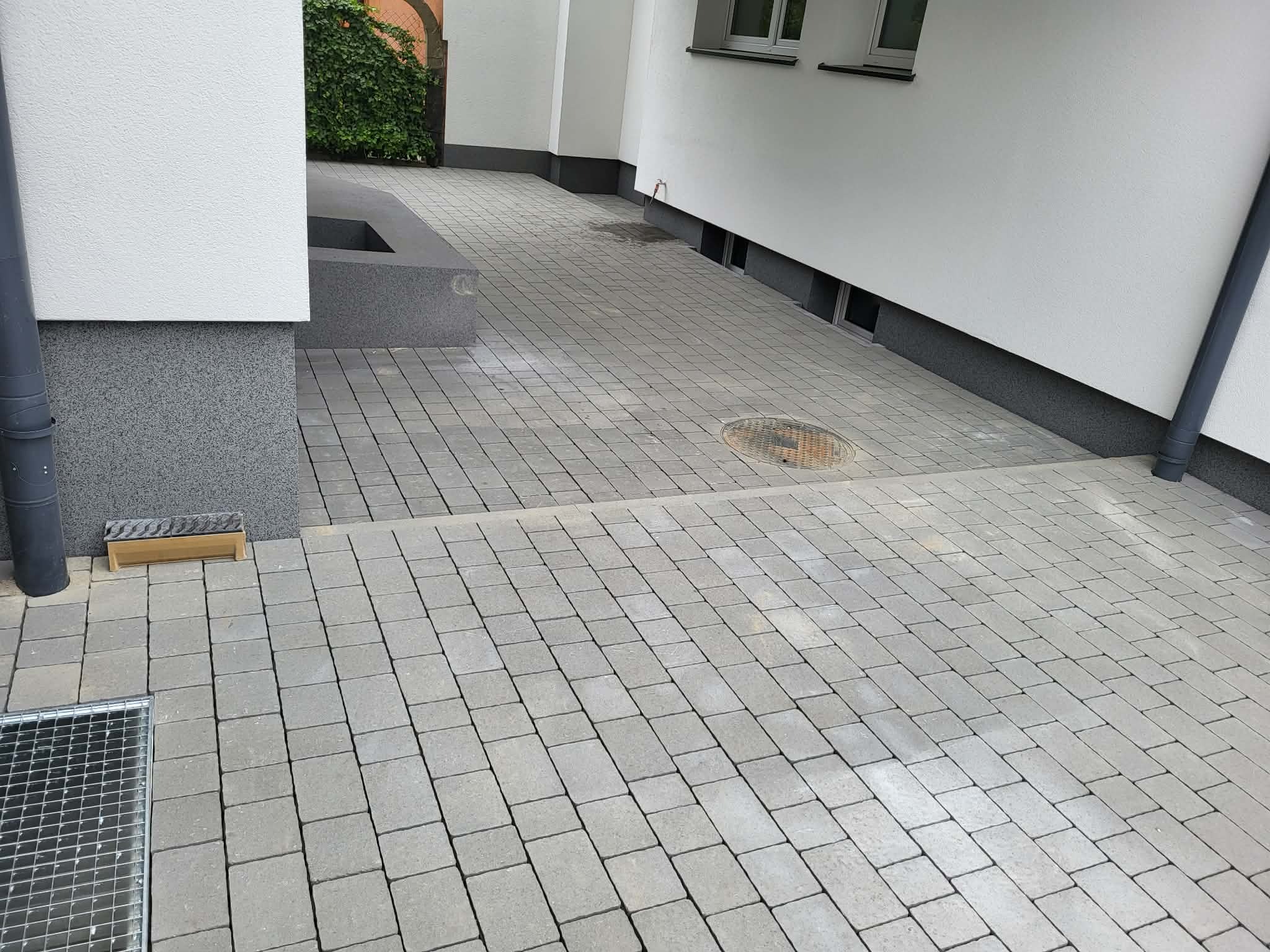 Szara kostka brukowa ułożona na podjeździe, widoczny fragment budynku z rurą spustową i kratką ściekową. Ujęcie z niskiej perspektywy podkreśla regularność wzoru.
