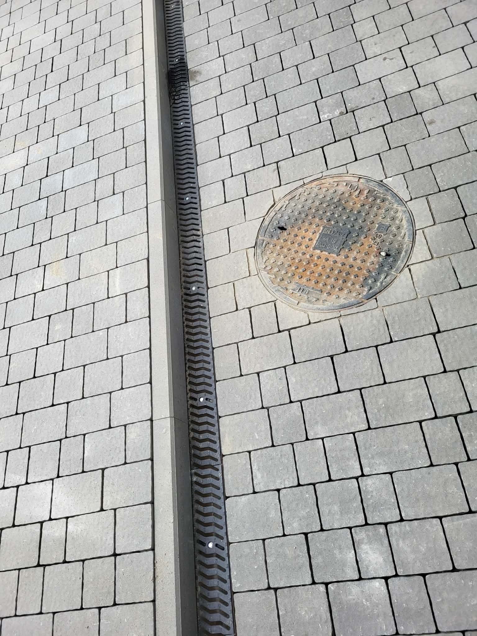 Szara kostka brukowa ułożona w regularny wzór, z liniowym odpływem wody i studzienką kanalizacyjną. Widok z góry, oświetlenie naturalne.