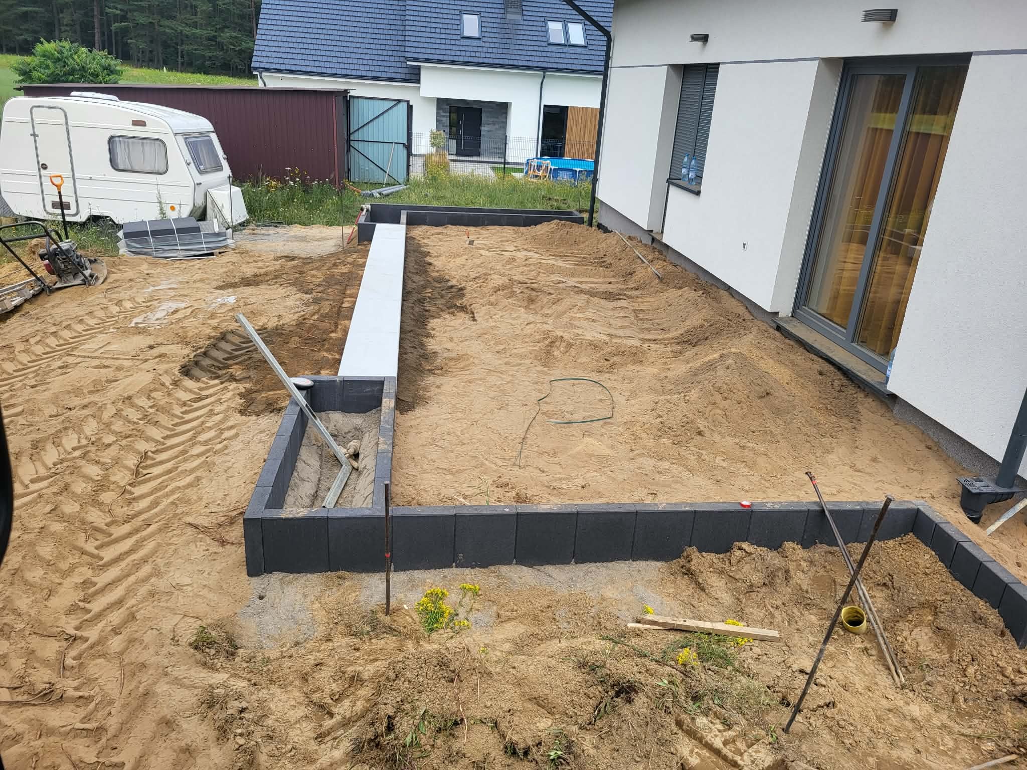 Przygotowanie terenu pod kostkę brukową: obrzeża z kostki, piasek, widoczne ślady opon i narzędzia. W tle dom i przyczepa kempingowa. Prace w toku.