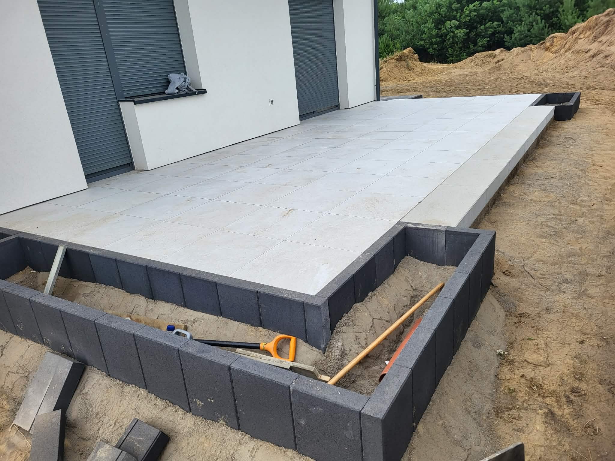 Jasne płytki tarasowe przy domu, obrzeża z ciemnej kostki. W wykopie narzędzia: łopata, poziomica. W tle usypana ziemia i zieleń. Etap prac brukarskich.