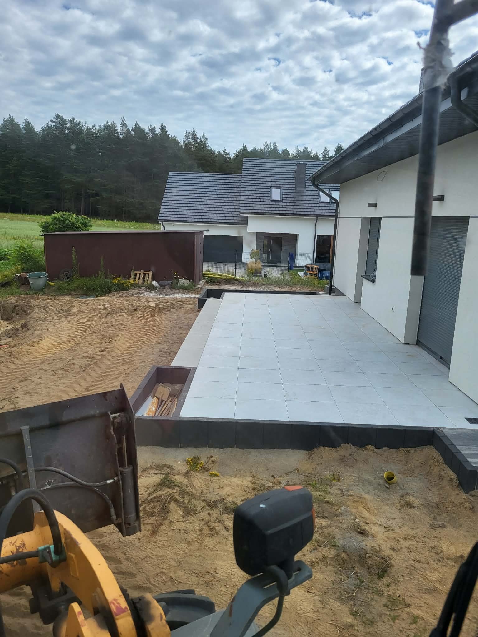 Jasne płytki tarasowe przy nowoczesnym domu, widok z kabiny koparki. Przygotowany teren pod dalsze prace, widoczne narzędzia i piasek. Częściowo zachmurzone niebo.
