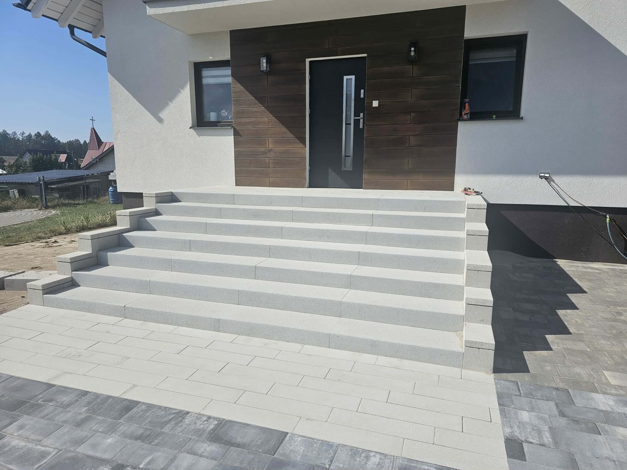 Nowoczesne schody wejściowe z jasnoszarej kostki brukowej prowadzące do domu z ciemnymi elementami elewacji i kostką w dwóch odcieniach szarości na podjeździe.