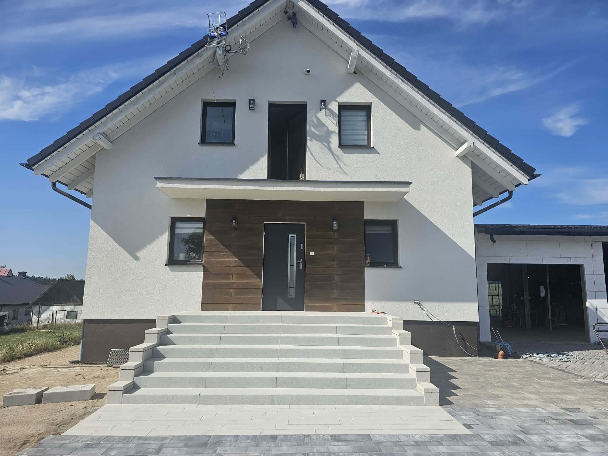 Nowoczesny dom jednorodzinny z białymi ścianami, brązowymi akcentami i schodami z jasnej kostki. Częściowo ułożona kostka brukowa na podjeździe. Błękitne niebo w tle.