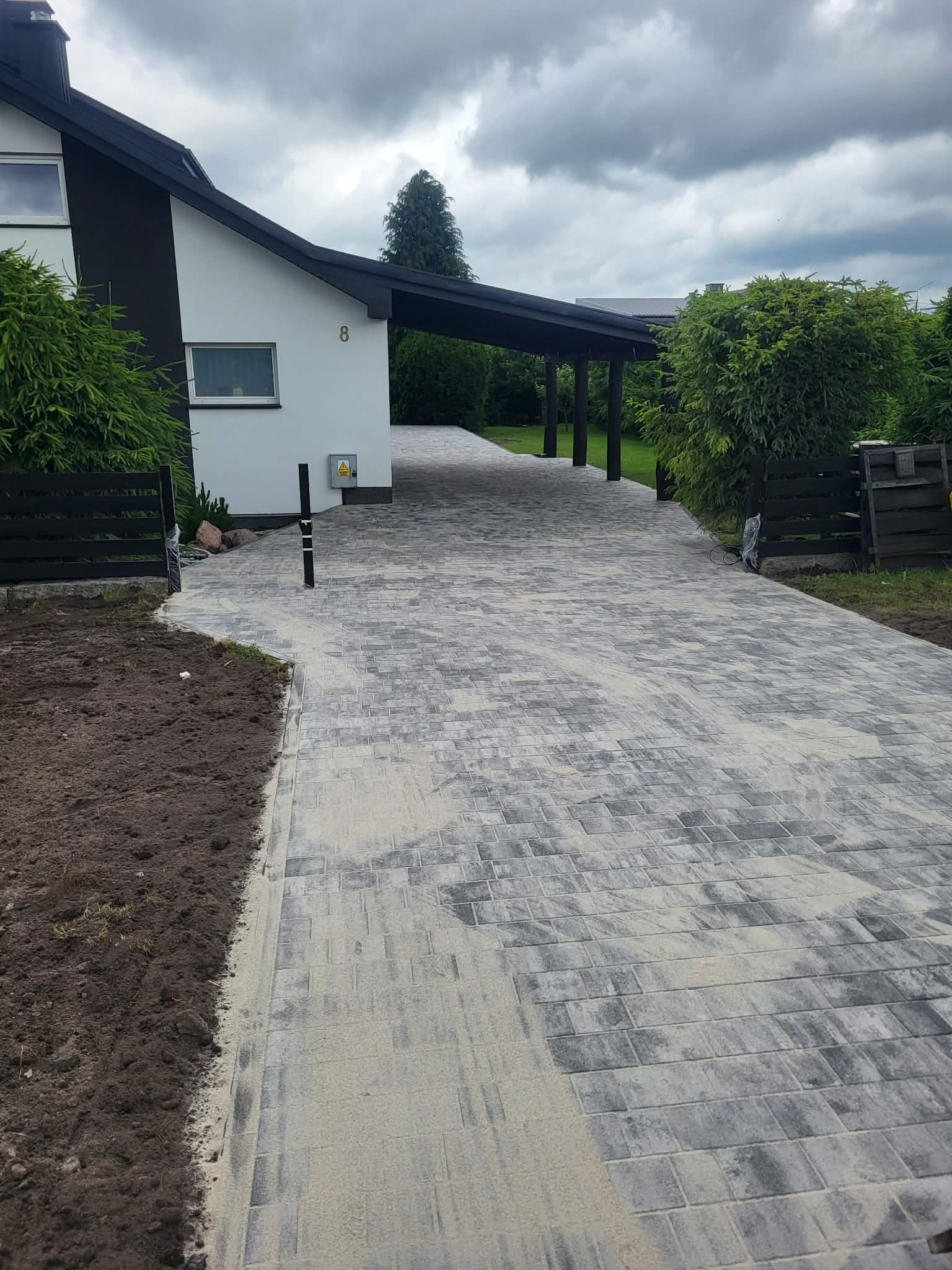 Nowa, szara kostka brukowa prowadząca do domu z numerem 8, widok z perspektywy. Świeżo ułożona nawierzchnia z piaskiem, obok rabata z ziemią.
