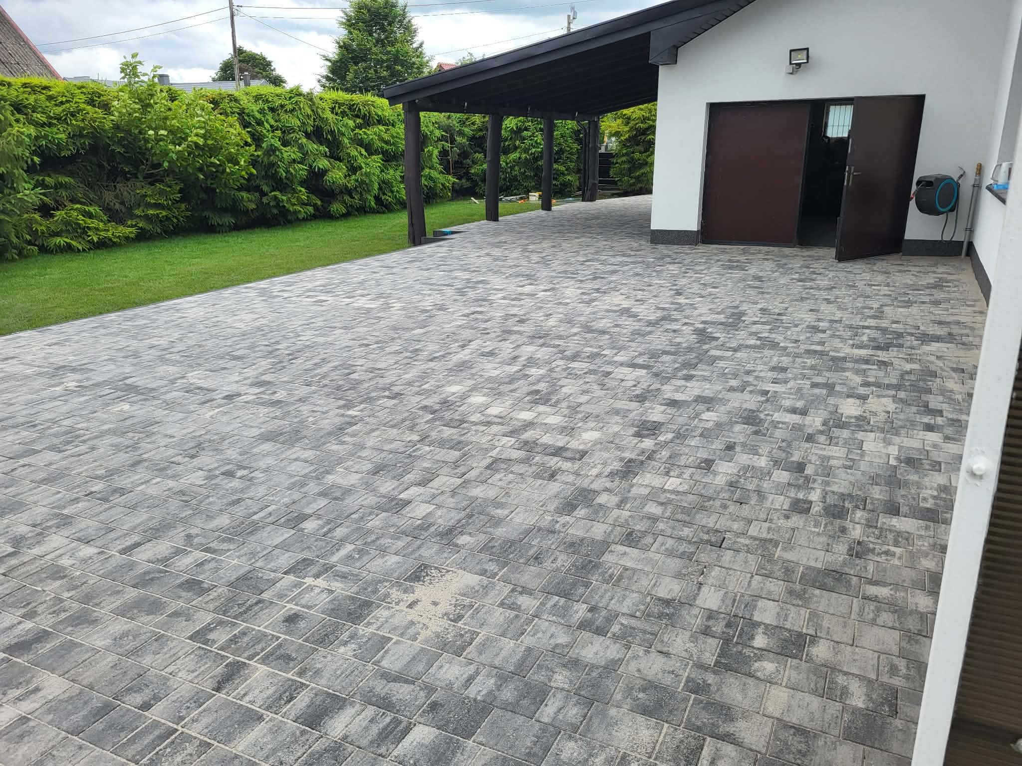 Ułożona szara kostka brukowa na podjeździe z zadaszeniem i garażem. Widoczny trawnik i krzewy. Kostka ułożona w regularny wzór, lekko zabrudzona piaskiem.