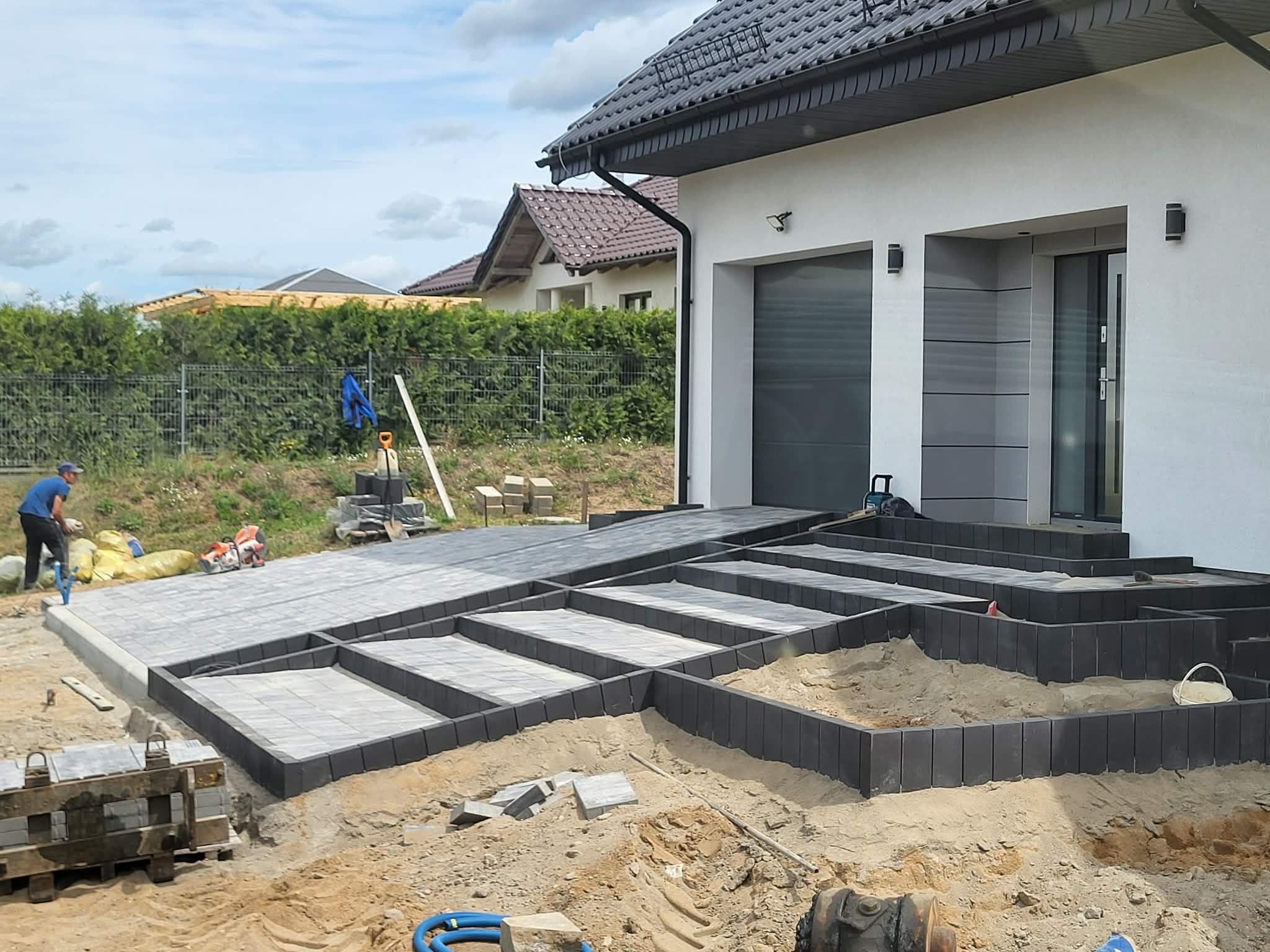 Etap prac: układanie kostki brukowej na podjeździe i schodach wejściowych do domu. Widoczne narzędzia i materiały budowlane. Prace brukarskie w toku.