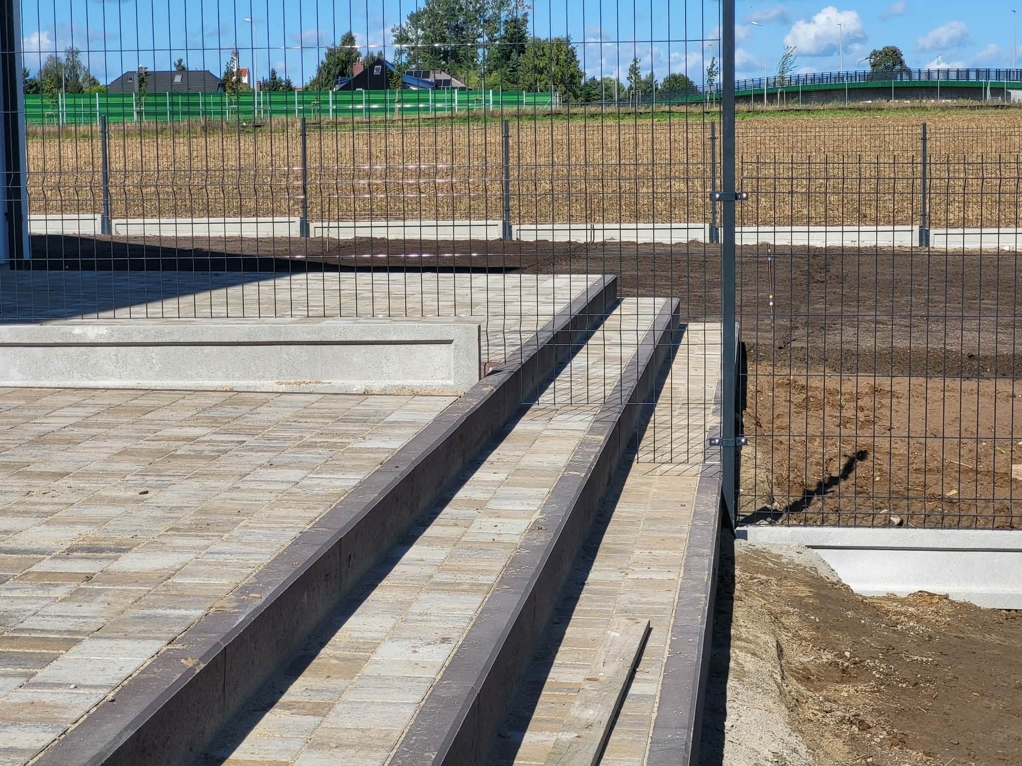 Schody z kostki brukowej z obrzeżami, prowadzące do metalowego ogrodzenia panelowego na tle zieleni i błękitnego nieba. Widoczne elementy krajobrazu.