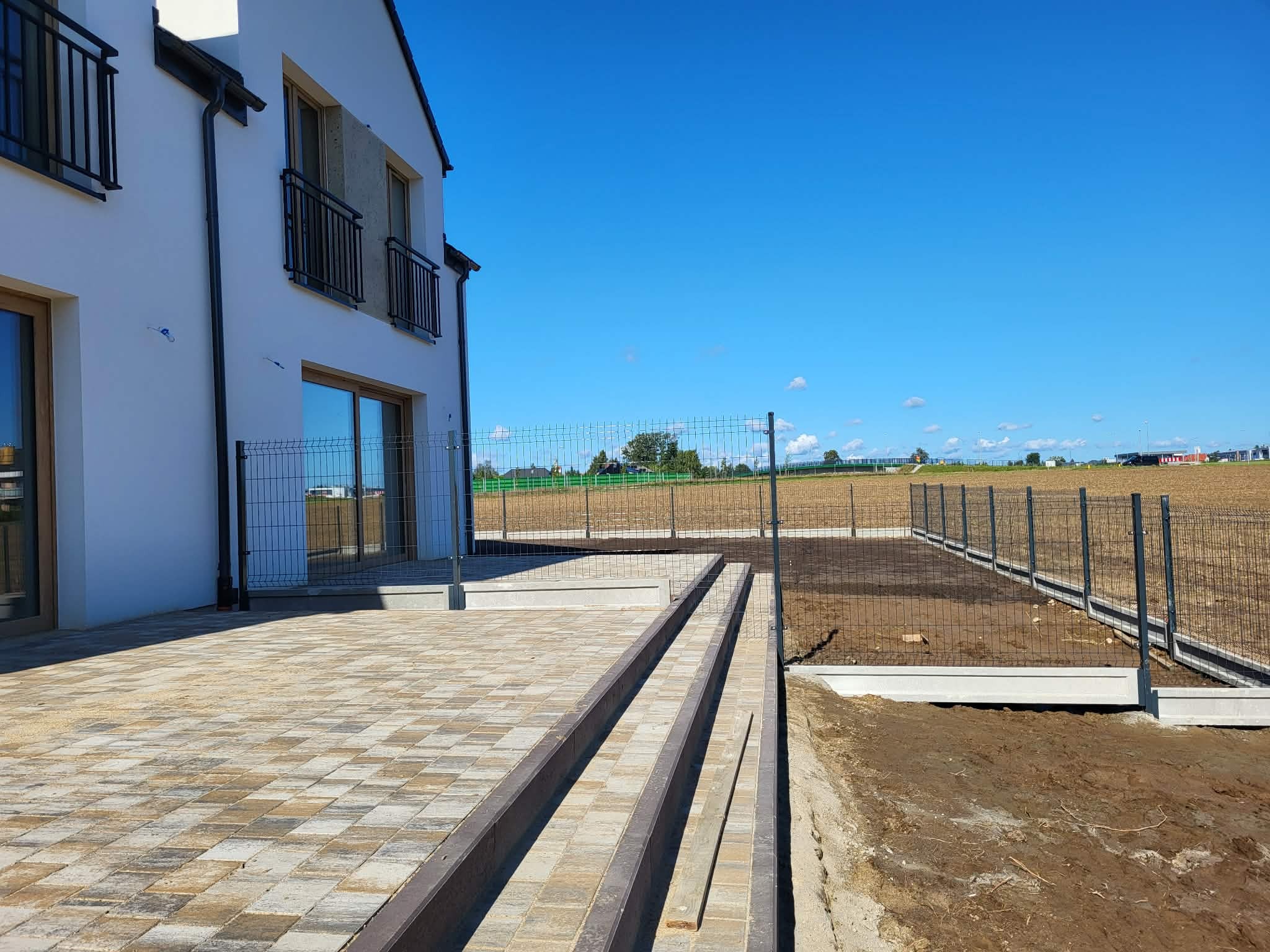 Nowoczesny dom z wybrukowanym tarasem i ogrodzeniem panelowym na tle błękitnego nieba. Widoczne schody z kostki brukowej i przygotowany teren pod trawnik.