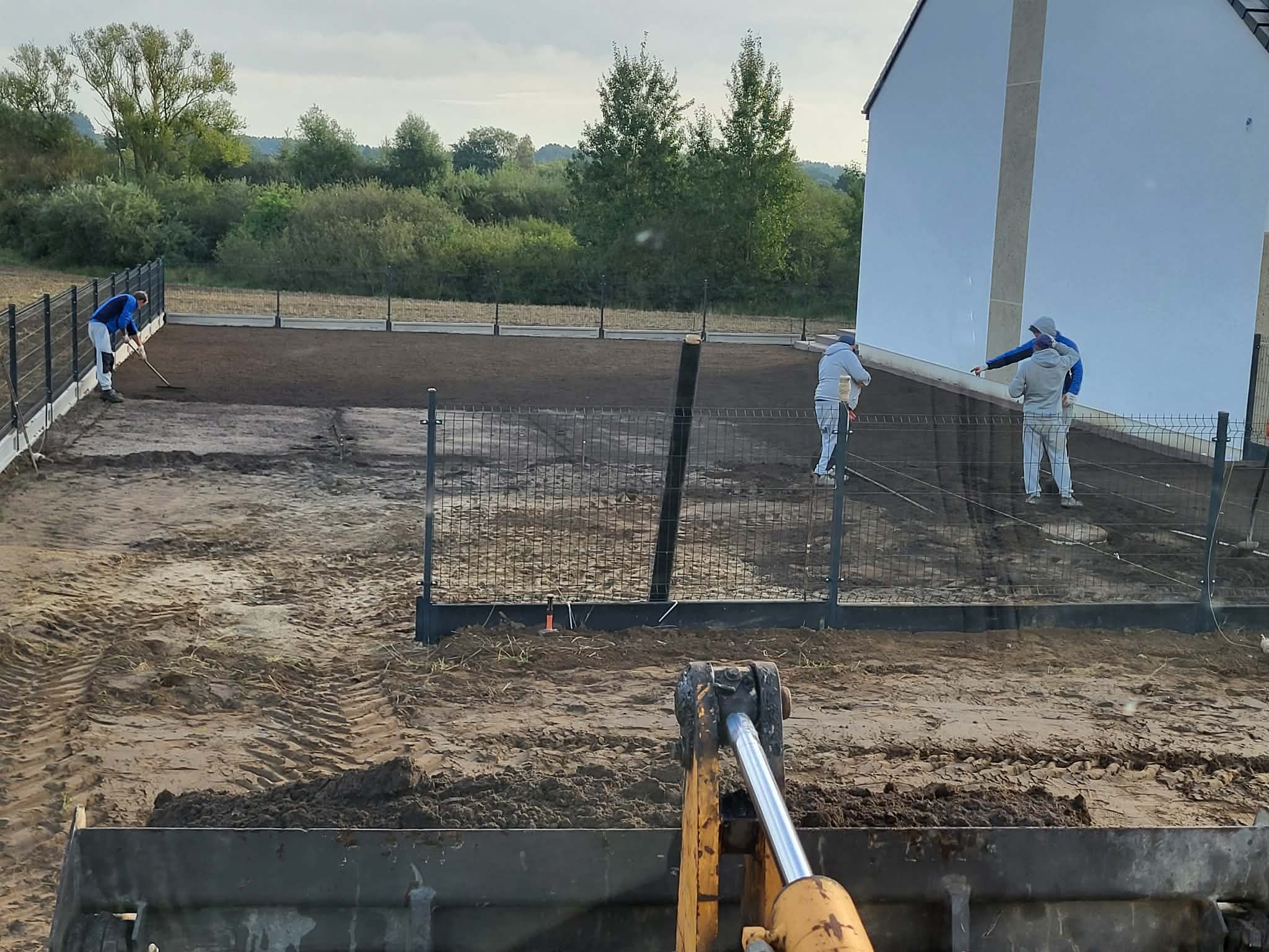 Niwelacja terenu pod budowę domu jednorodzinnego. Widoczny operator koparki, pracownicy wyrównujący teren, ogrodzenie panelowe i budynek w tle.