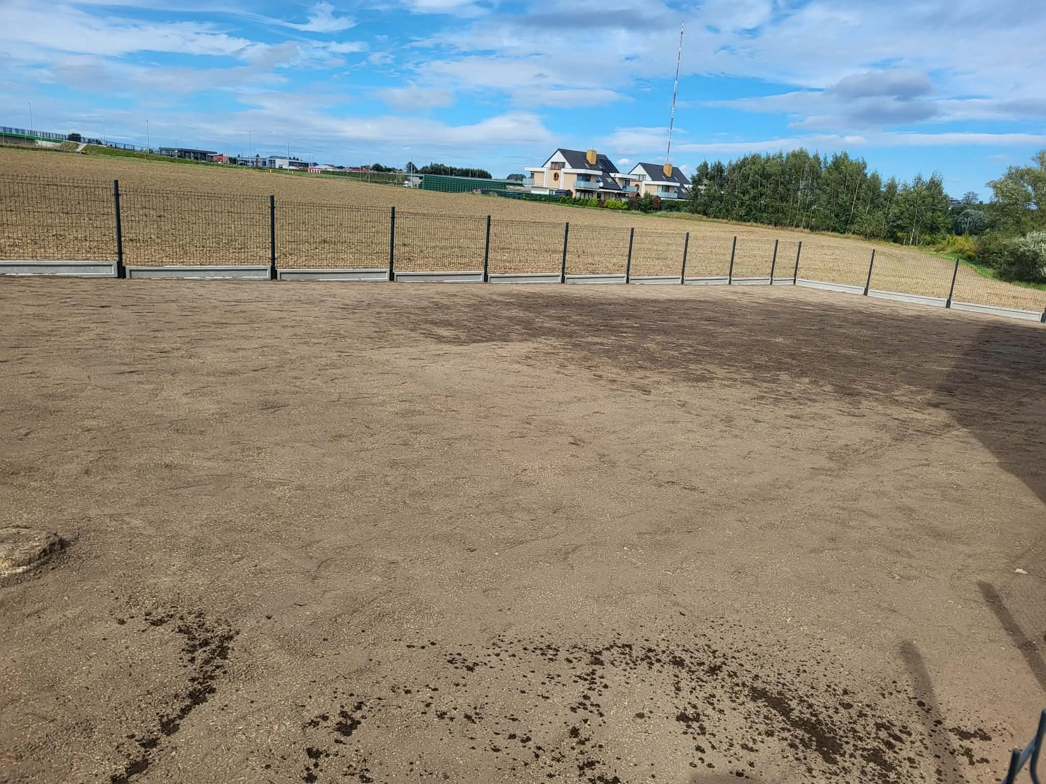 Nowoczesne ogrodzenie panelowe z podmurówką na wyrównanym terenie. W tle domy jednorodzinne i błękitne niebo z chmurami. Widok z poziomu gruntu.
