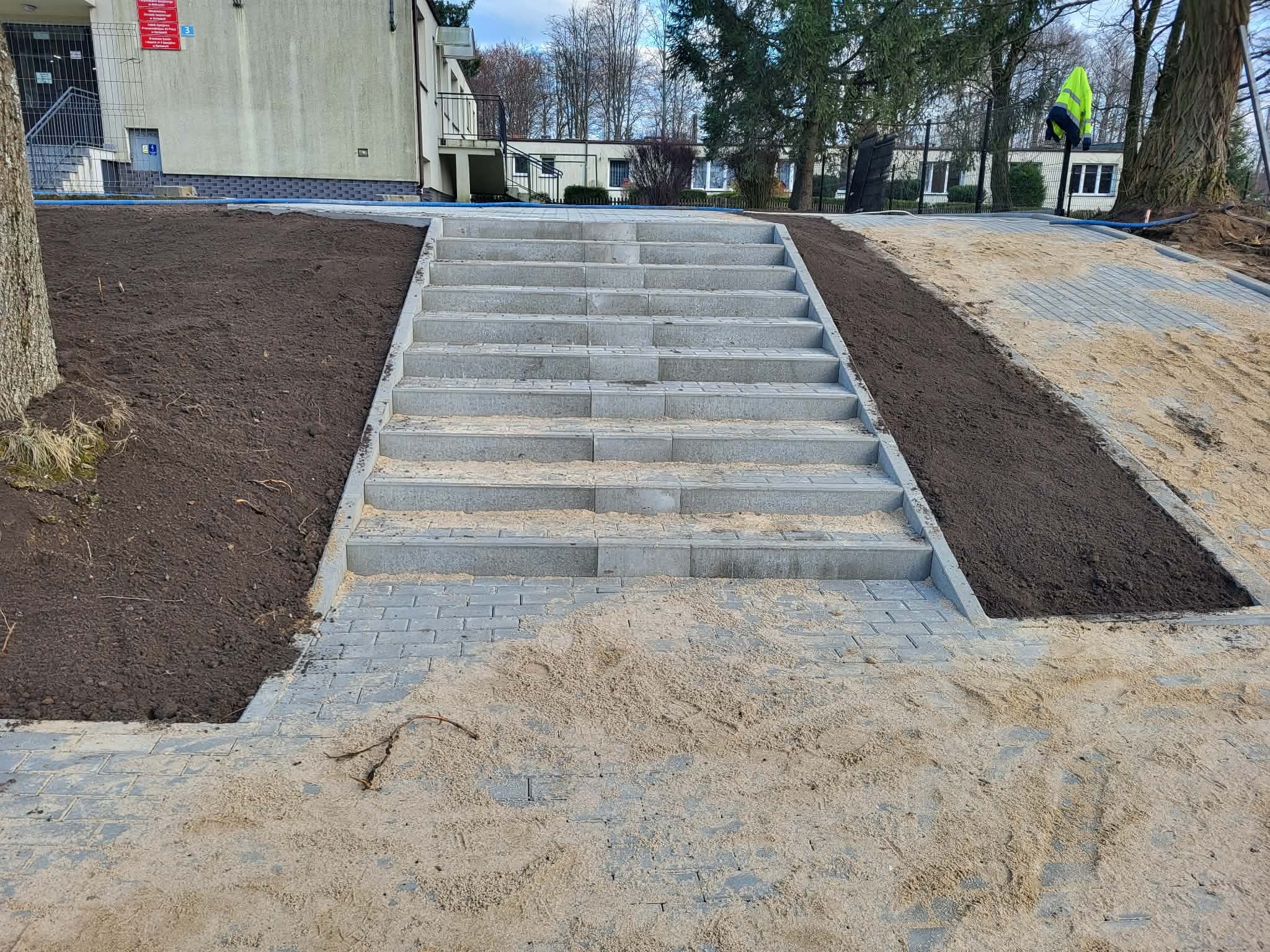 Schody z kostki brukowej prowadzące w górę, otoczone ziemią i piaskiem. Widoczne budynki w tle i drzewa. Ujęcie z dołu, podkreślające perspektywę schodów.