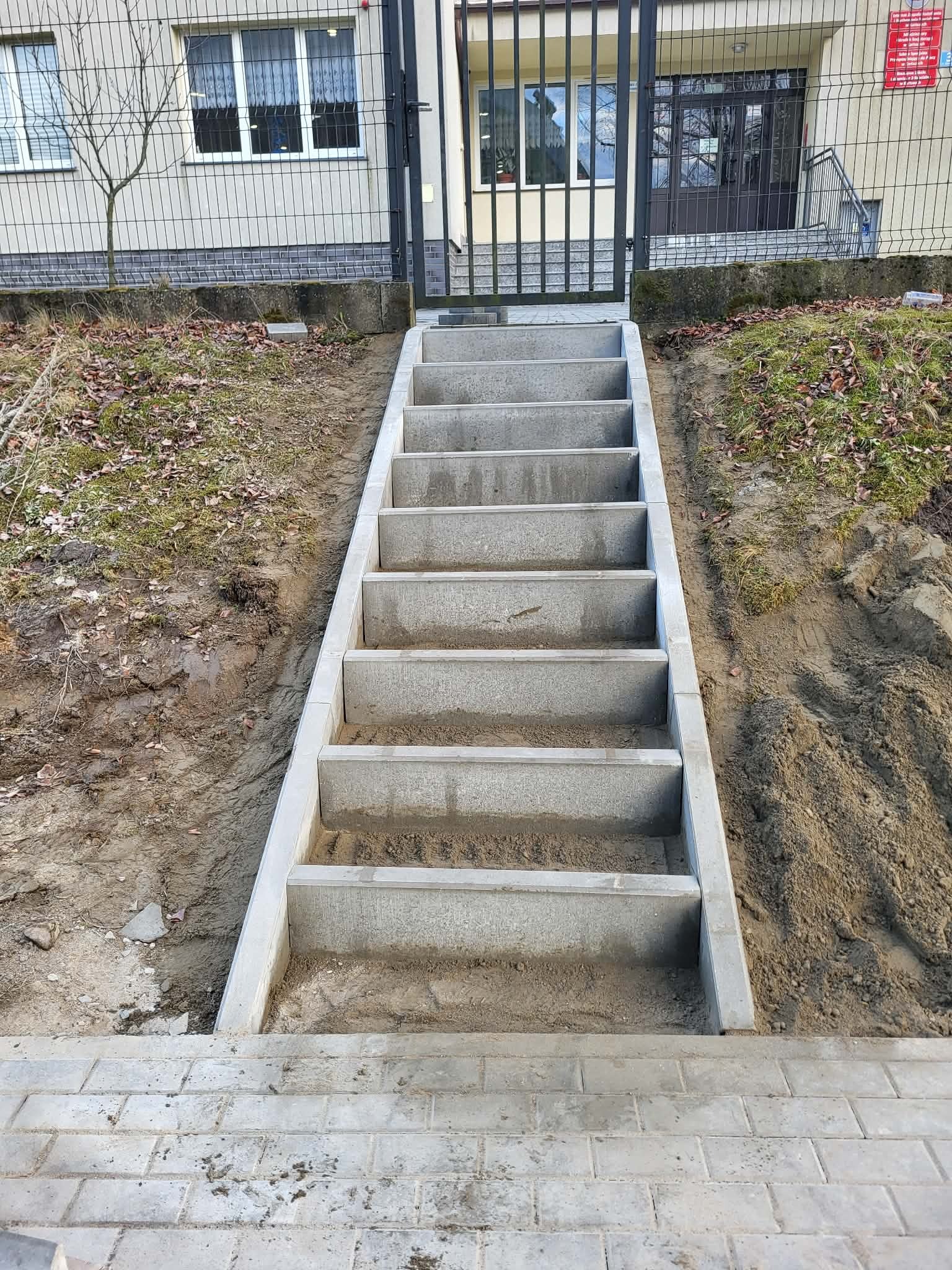 Betonowe schody prowadzące do budynku w Popowie, otoczone ziemią i kostką brukową. Widoczne ogrodzenie i brama wejściowa. Ujęcie z dołu, podkreślające solidność konstrukcji.
