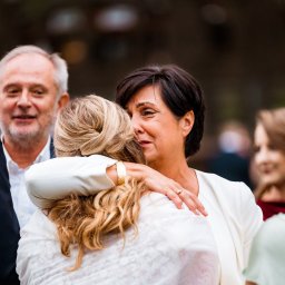 Fotoopowieści Wojciech Gontarz - Uścisk kobiety w bieli z blondynką na tle uśmiechniętego mężczyzny i innych osób. Ciepłe oświetlenie, emocjonalna scena.
