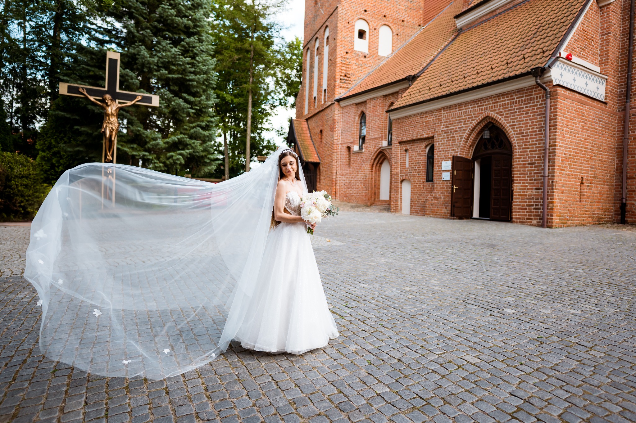 Panna młoda w zwiewnej sukni i długim welonie z bukietem stoi na tle ceglanego kościoła i krzyża. Romantyczna sceneria, kamienna nawierzchnia.
