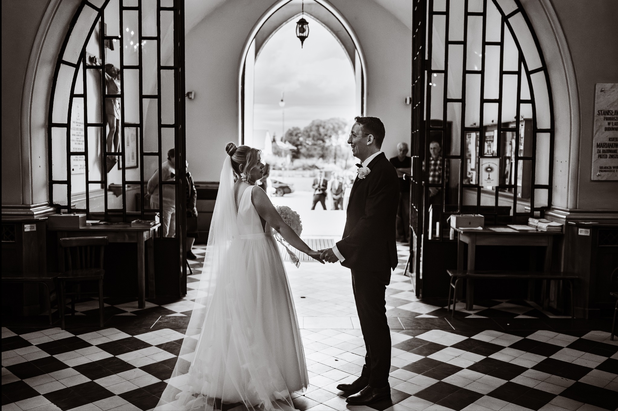 Czarno-białe zdjęcie pary młodej trzymającej się za ręce w kościele, z widokiem na zewnątrz przez otwarte drzwi. W tle widoczni goście i elementy architektoniczne.