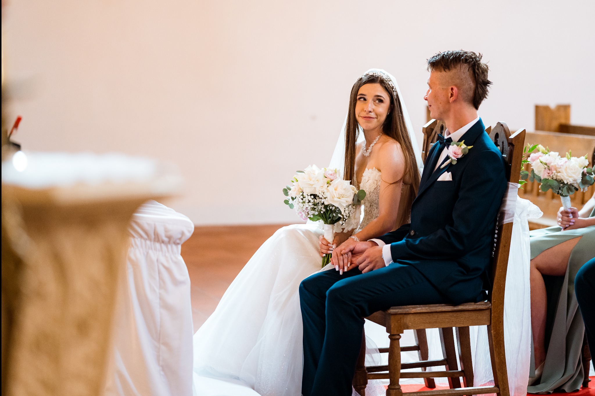 Para młoda siedzi obok siebie podczas ceremonii ślubnej. Panna młoda trzyma bukiet białych kwiatów, a pan młody ma ciemny garnitur z butonierką.