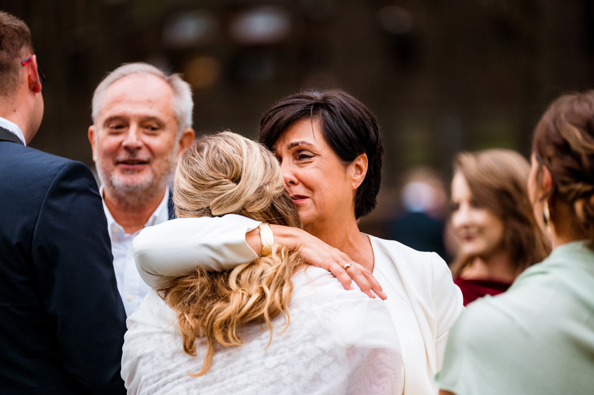 Uścisk kobiety w bieli z blondynką na tle uśmiechniętego mężczyzny i innych osób. Ciepłe oświetlenie, emocjonalna scena.