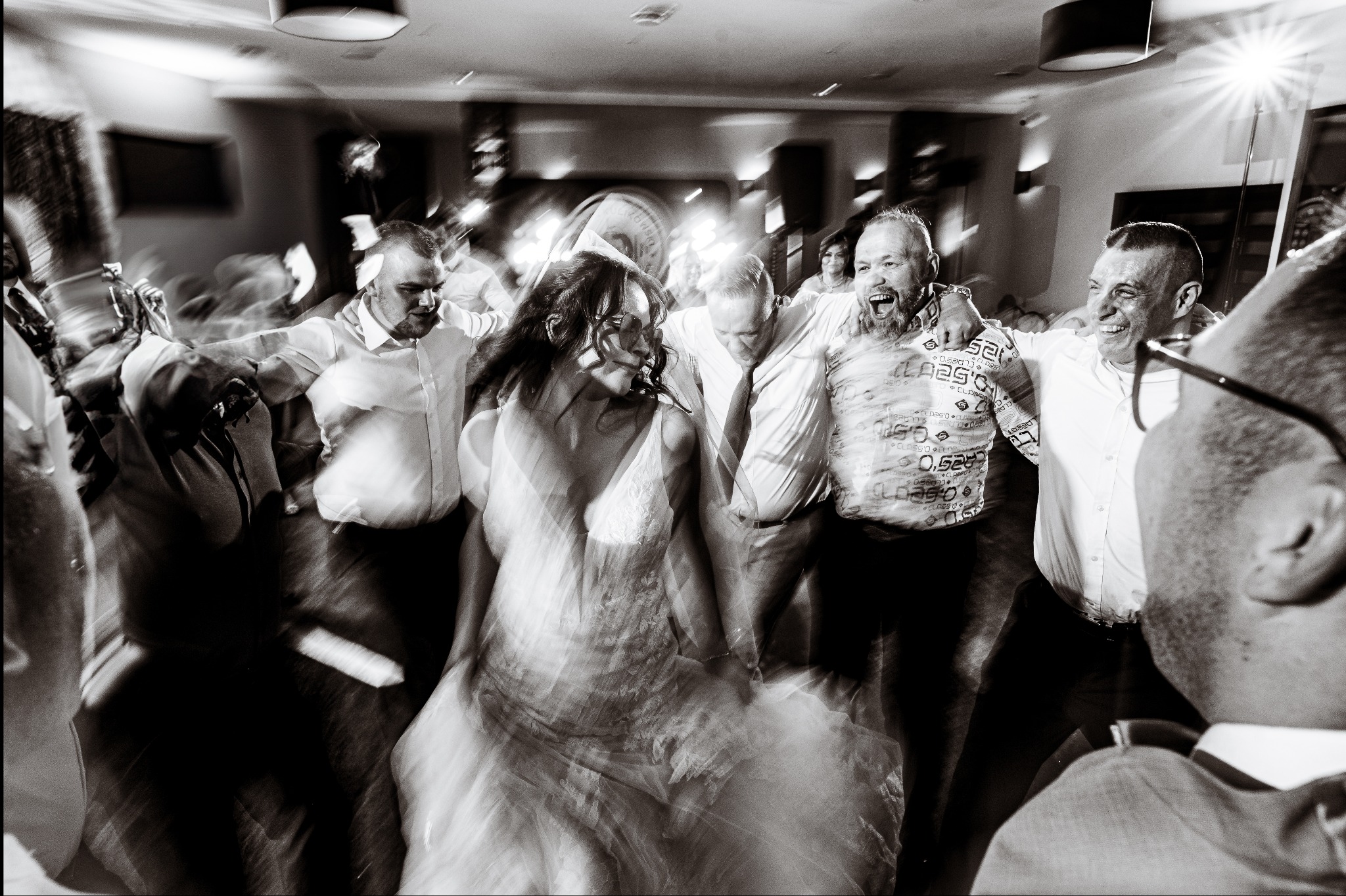 Rozmazane, czarno-białe zdjęcie z wesela: panna młoda w tańcu z gośćmi, dynamiczne ujęcie z efektem ruchu, radosne twarze, światło reflektorów.
